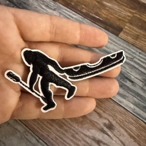 May include: A black embroidered patch featuring a silhouette of a creature resembling Bigfoot or a yeti, depicted in a walking motion while carrying a long, thin pole with a small attachment on one end and a larger, curved object on the other.