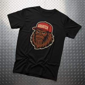 May include: Black t-shirt featuring a cartoon illustration of a Bigfoot wearing a red baseball cap. The cap has the word "Squatch" printed on it in white. The Bigfoot has brown fur and a serious expression.