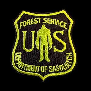 May include: A black and yellow embroidered patch with the text "FOREST SERVICE" and "DEPARTMENT OF SASQUATCH". The patch features a yellow silhouette of Bigfoot within a shield design.