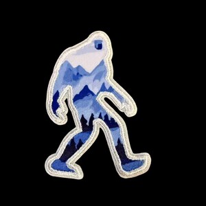 May include: A Bigfoot patch with a white border. The interior features a blue and white mountain scene. The patch is in the shape of the legendary creature, with a walking pose. The background is black.