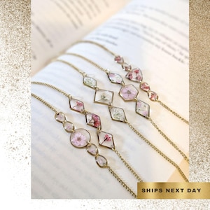 May include: Several gold-toned chain bracelets with clear geometric pendants containing dried flowers in shades of pink and white. The bracelets are displayed on an open book. The text "SHIPS NEXT DAY" is visible.