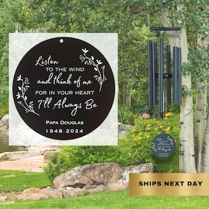 May include: Black metal wind chime with a round, black metal plate that reads "Listen to the wind and think of me for in your heart I'll always be Papa Douglas 1948-2024". The wind chime is hanging from a tree branch in a garden setting.