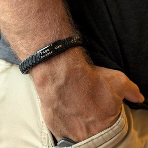 Personalized Black Leather Bracelet for Men – Engraved Magnetic Clasp | Custom Gift for Husband, Boyfriend or Dad, Custom Father's Day name. Handcrafted from genuine black braided leather with a sleek engraved black clasp, this personalized bracelet is rugged, refined, and ridiculously handsome. Close-up image of a man's wrist wearing a black braided leather bracelet engraved with the words "Papa Est. 2024" and "Liam". The bracelet is personalized and designed as a gift for new fathers