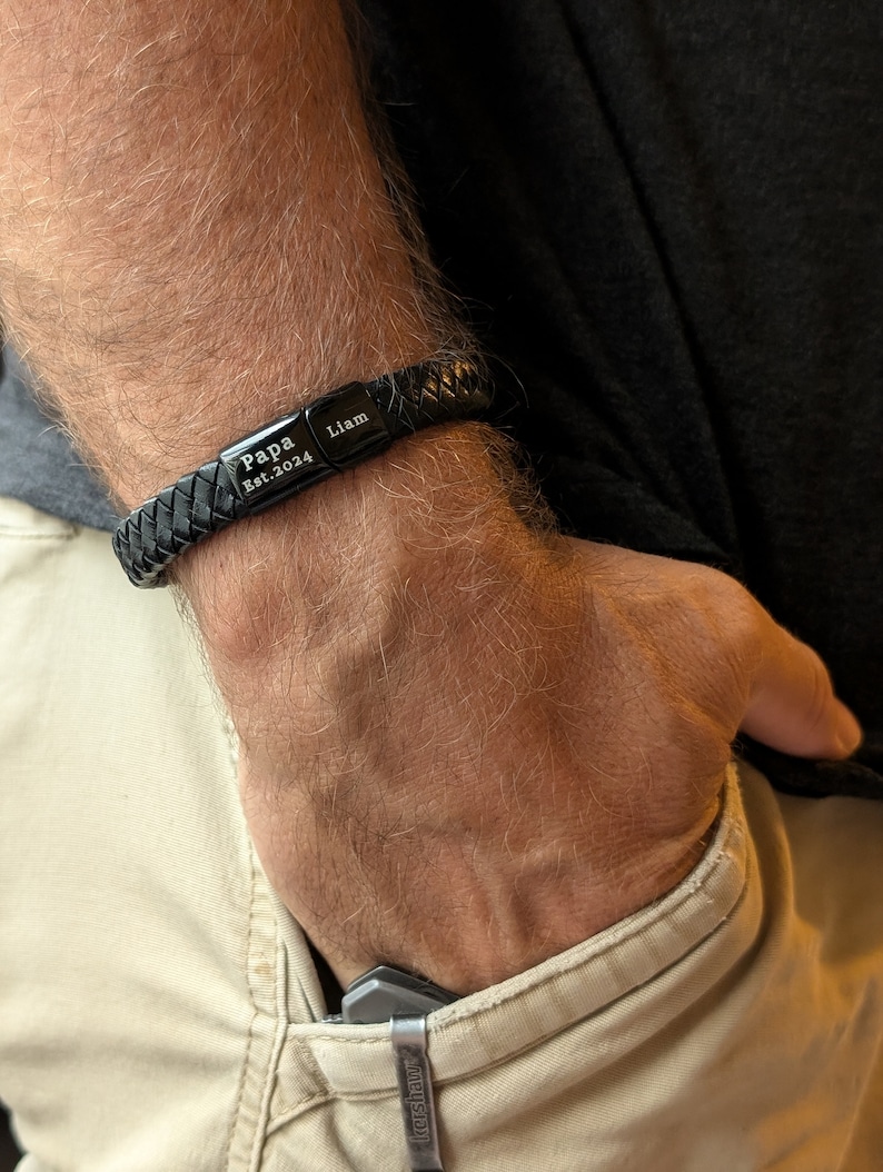 Personalized Black Leather Bracelet for Men – Engraved Magnetic Clasp | Custom Gift for Husband, Boyfriend or Dad, Custom Father's Day name. Handcrafted from genuine black braided leather with a sleek engraved black clasp, this personalized bracelet is rugged, refined, and ridiculously handsome. Close-up image of a man's wrist wearing a black braided leather bracelet engraved with the words "Papa Est. 2024" and "Liam". The bracelet is personalized and designed as a gift for new fathers