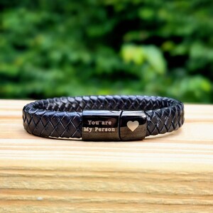 Personalized Black Leather Bracelet for Men – Engraved Magnetic Clasp | Custom Gift for Husband, Boyfriend or Dad, Custom Father's Day name.  Black braided leather bracelet with engraved black metal plates that read “You are My Person” and feature a small heart symbol. Displayed on a wooden surface with a blurred green outdoor background. A meaningful, unisex gift perfect for anniversaries, best friends, or loved ones.