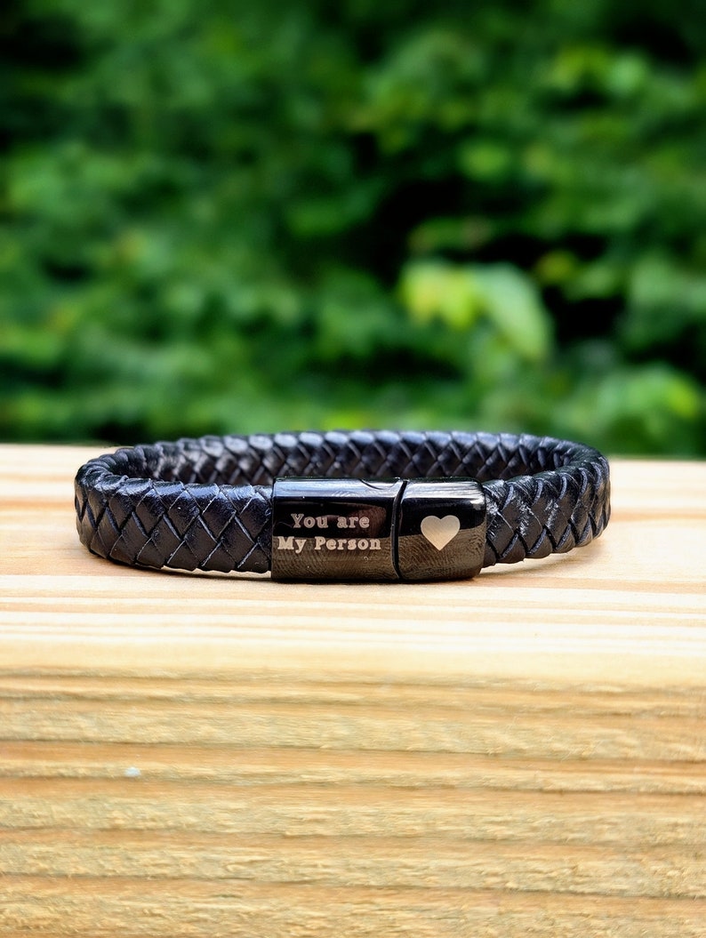 Personalized Black Leather Bracelet for Men – Engraved Magnetic Clasp | Custom Gift for Husband, Boyfriend or Dad, Custom Father's Day name.  Black braided leather bracelet with engraved black metal plates that read “You are My Person” and feature a small heart symbol. Displayed on a wooden surface with a blurred green outdoor background. A meaningful, unisex gift perfect for anniversaries, best friends, or loved ones.