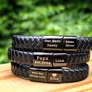Personalized Black Leather Bracelet for Men – Engraved Magnetic Clasp | Custom Gift for Husband, Boyfriend or Dad, Custom Father's Day name. Stack of three custom engraved black braided leather bracelets, each with personalized metal plates. Engravings include phrases like “Our Hero Daddy,” “Papa Est. 2024,” “Have Fun Always Love,” and names like Emma, Oliver, Liam, and Ellie with a heart symbol. Displayed outdoors on a wooden surface with a green background, a heartfelt, masculine gift.