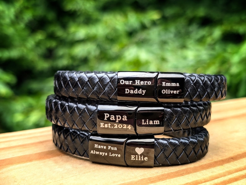 Personalized Black Leather Bracelet for Men – Engraved Magnetic Clasp | Custom Gift for Husband, Boyfriend or Dad, Custom Father's Day name. Stack of three custom engraved black braided leather bracelets, each with personalized metal plates. Engravings include phrases like “Our Hero Daddy,” “Papa Est. 2024,” “Have Fun Always Love,” and names like Emma, Oliver, Liam, and Ellie with a heart symbol. Displayed outdoors on a wooden surface with a green background, a heartfelt, masculine gift.