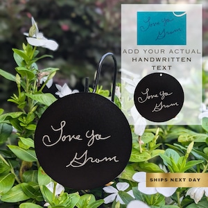 May include: Black circular metal sign with a white inscription that reads "Love you Grom". The sign is hanging from a metal hook.
