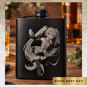 May include: A matte black metal flask featuring a laser-engraved illustration of an anime-style character with long flowing hair, wearing a sleeveless top and arm warmers. The flask has a hinged screw-top cap with a textured grip pattern.