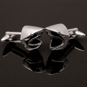 May include: A pair of silver-toned cufflinks, each shaped like a shark head with an open mouth revealing teeth. The cufflinks are highly polished and reflect the dark surface they rest upon. The design is detailed and the overall aesthetic is modern.