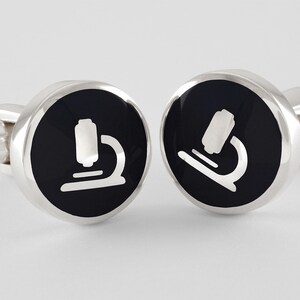 May include: A pair of silver cufflinks with black enamel and a white microscope design.