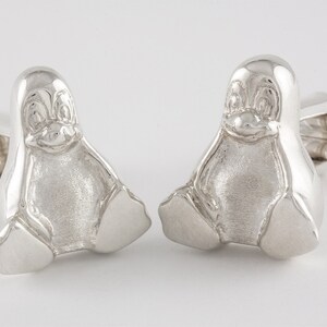 May include: A pair of silver cufflinks featuring a penguin design. The penguins are sitting with their wings folded and have a detailed face.