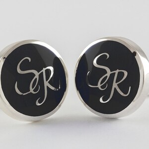 May include: A pair of silver cufflinks with black enamel and the initials "SR" engraved in a script font.
