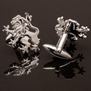 May include: A pair of silver cufflinks featuring a lion design. The cufflinks are shown on a black surface.