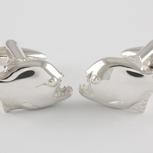 May include: A pair of silver cufflinks shaped like Piranha fish. The fish are facing each other with their mouths open.
