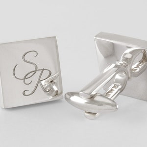 May include: A pair of square silver cufflinks. One cufflink features the engraved initials "SR" in a cursive font. The other cufflink is angled to show the clasp and the manufacturer's mark. The cufflinks are set against a white background.
