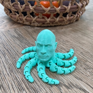 May include: A light teal 3D-printed octopus figurine with a human face. The octopus has articulated tentacles and is displayed on a wooden surface. A woven basket filled with fruit is in the background.