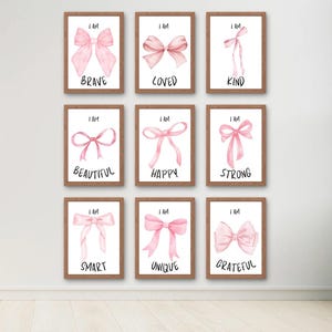 May include: Nine framed art prints with watercolor illustrations of pink bows. Each print features the words "I AM" above a positive affirmation: Brave, Loved, Kind, Beautiful, Happy, Strong, Smart, Unique, and Grateful. The frames are brown.