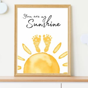 May include: A framed print with the text "You are my Sunshine" above a yellow watercolor sun design. The sun features two yellow footprints at the center. The frame is a light wood color.
