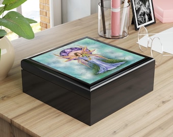 May include: A black wooden box with a hinged lid. The lid features a colorful illustration of a fairy with purple and gold wings, wearing a blue dress, set against a light blue and green background. The box is sitting on a wooden surface.