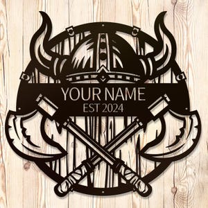 Puede incluir: Arte de pared de metal negro con un casco vikingo con cuernos, hachas cruzadas y una placa de nombre personalizada con el texto "YOUR NAME EST 2024".