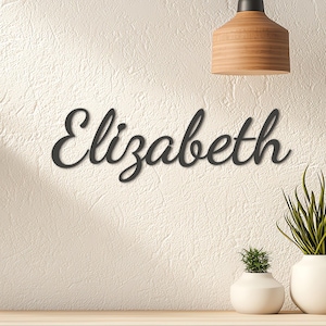 May include: A black metal sign displaying the name "Elizabeth" in a cursive font. The sign is mounted on a textured, off-white wall. Decorative elements include a wooden lamp and white vases with plants.