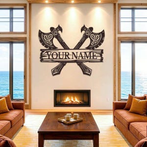 Op de afbeelding: Een donkerbruin metalen wandbord met twee gekruiste bijlen met ingewikkelde uitgesneden patronen. Een horizontale rechthoekige banner is over het midden van de bijlen geplaatst en toont de tekst YOUR NAME in dikke hoofdletters.