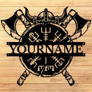 Puede incluir: Arte de pared de metal negro con un casco vikingo, hachas y un escudo con runas. El texto "YOURNAME" se muestra en el escudo. La obra de arte está sobre un fondo de madera.