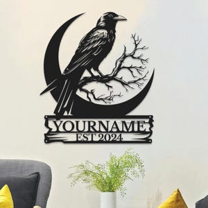 May include: Black metal wall art featuring a raven perched on a branch within a crescent moon. The design includes the text "YOURNAME EST 2024" in a decorative banner. The artwork is mounted on a white wall.