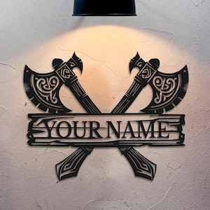 Op de afbeelding: Zwarte metalen wanddecoratie met twee gekruiste bijlen met ingewikkelde ontwerpen en een gepersonaliseerd naamplaatje met de tekst "YOUR NAME".