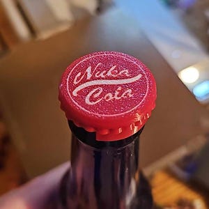 Puede incluir: Tapa de botella roja con un logotipo blanco que dice "Nuka Cola" en una fuente estilizada.