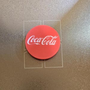 Può includere: Un logo Coca-Cola rotondo e rosso. Il logo in scrittura bianca è centrato sullo sfondo rosso. L'immagine è su una superficie metallica strutturata.