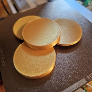 May include: Four round, gold-colored coasters on a black surface. The coasters are smooth and have a slight sheen.