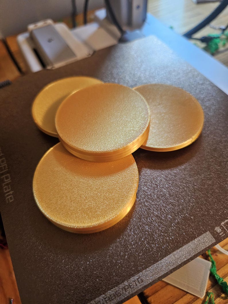 May include: Four round, gold-colored coasters on a black surface. The coasters are smooth and have a slight sheen.