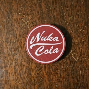 Può includere: Un adesivo circolare rosso e bianco con il testo "Nuka Cola" in un font bianco corsivo.