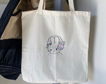 May include: Cream-colored tote bag with a black line drawing of a bunny wearing headphones. The headphones have colorful accents. The bag is hanging on a metal bar next to a dark blue jacket.