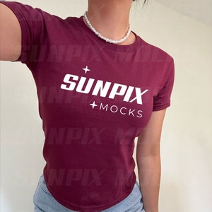 Può includere: Una maglietta bordeaux con la scritta "SUNPIX + MOCKS" in bianco. La maglietta è abbinata a pantaloncini di jeans azzurri. Una collana di perle è indossata intorno al collo.