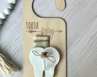 May include: Wooden door hanger with the text "TOOTH fairy" and "PLEASE STOP HERE". Features a white tooth shape with a small glass bottle and a slot for money. Includes a hanging loop.