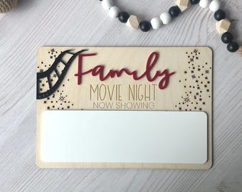May include: Wooden sign with the words "Family Movie Night" in red script and block letters. A black film reel graphic is on the left. The sign has a white rectangular area for personalization. Decorative stars are scattered around the sign.