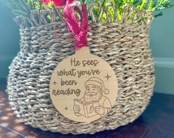 May include: A round wooden Christmas ornament with the text "He sees what you've been reading" and a Santa Claus illustration. The ornament hangs from a red ribbon in front of a woven basket filled with greenery.