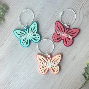 Könnte beinhalten: Drei schmetterlingsförmige Weinanhänger in Türkis, Rosa und Pfirsich. Jeder Anhänger hat einen weißen Drahtring und einen Namen in weißer Schrift: Ophelia, Angelina und Ruby. Die Anhänger liegen auf einer hellen Holzmaserung.