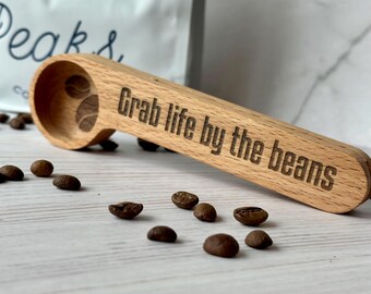 May include: A wooden coffee scoop with the phrase "Grab life by the beans" engraved on the handle. The scoop features a coffee bean design and is surrounded by coffee beans on a white wooden surface.
