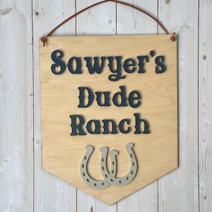 May include: Wooden sign with the text "Sawyer's Dude Ranch" in dark blue letters, with two gray horseshoes below. The sign is light brown with a leather hanging strap.