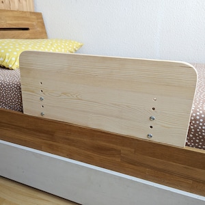 Può includere: Sponda del letto in legno chiaro fissata a un telaio del letto. La sponda è rettangolare con angoli arrotondati e fissata con viti. Il letto ha un lenzuolo fantasia e un cuscino giallo a pois.