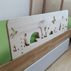 Puede incluir: Un barandal de cama de madera clara con un diseño recortado de un erizo, mariposas y plantas. El barandal está unido a un marco de cama de madera.