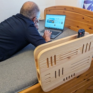Può includere: Un telaio per letto in legno con un tavolino laterale integrato e un porta telecomando. Il tavolino laterale ha un motivo geometrico di fessure ed è realizzato in legno chiaro. Una persona è sdraiata sul letto usando un laptop.