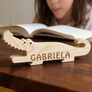 Peut inclure: Un porte-nom en forme d'alligator en bois avec le nom "GABRIELA" gravé sur le côté. L'alligator est de couleur claire et repose sur une surface en bois sombre. Le porte-nom est devant un livre ouvert.