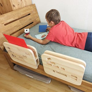 Könnte beinhalten: Ein hölzerner Bettrahmen mit einem Kopfteil und zwei Nachttischen. Die Nachttische haben ausgeschnittene Designs. Ein rotes Blatt und ein blaues Blatt liegen auf dem Bett. Eine Person liegt auf dem Bett und hält ein Spielzeug.