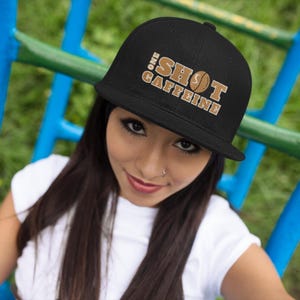 Può includere: Un cappello da baseball nero e marrone con il testo "One Shot Caffeine" in lettere dorate. Il cappello ha una visiera curva e una chiusura a scatto.